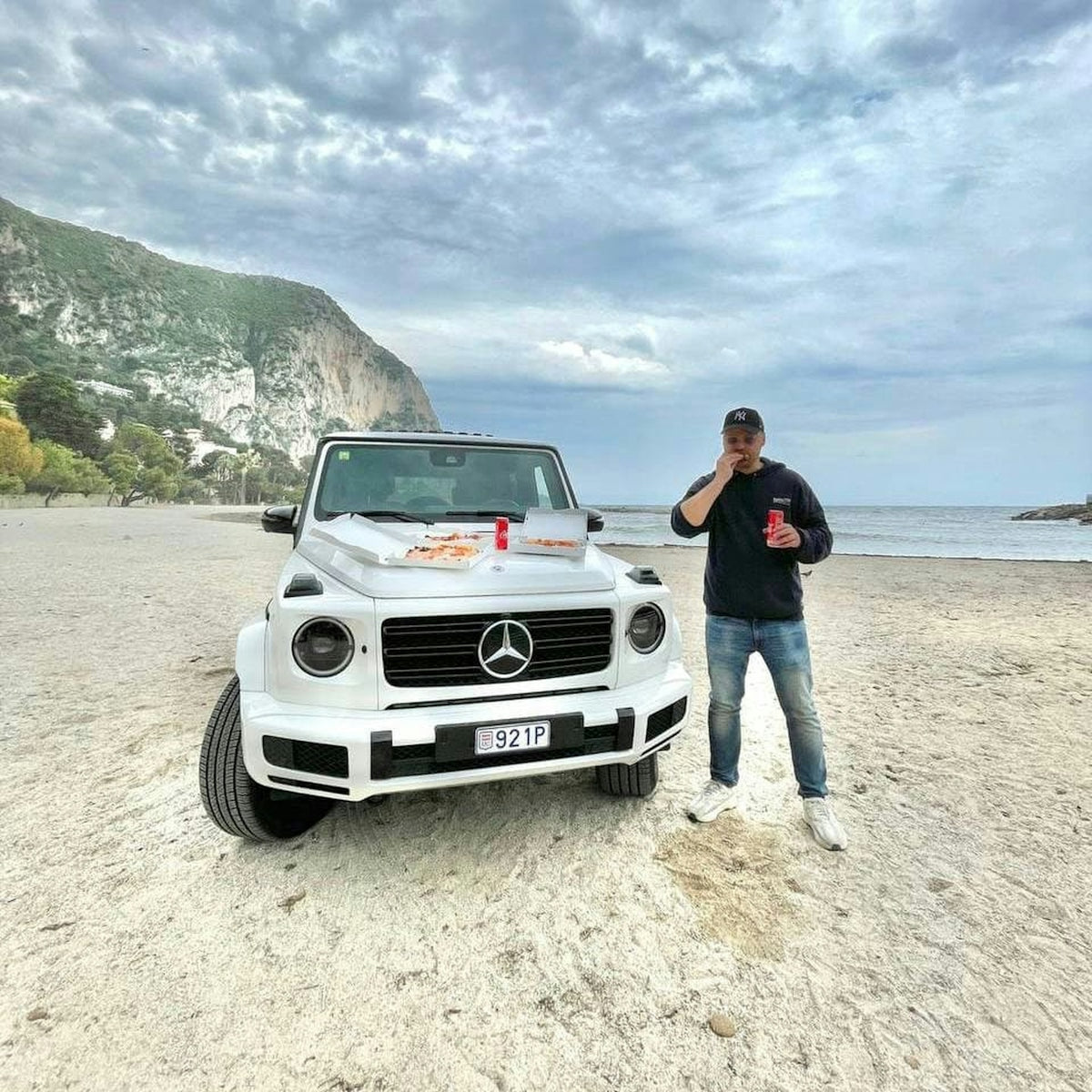 Man standing by white mercedes g-wagon on a sandy beach.