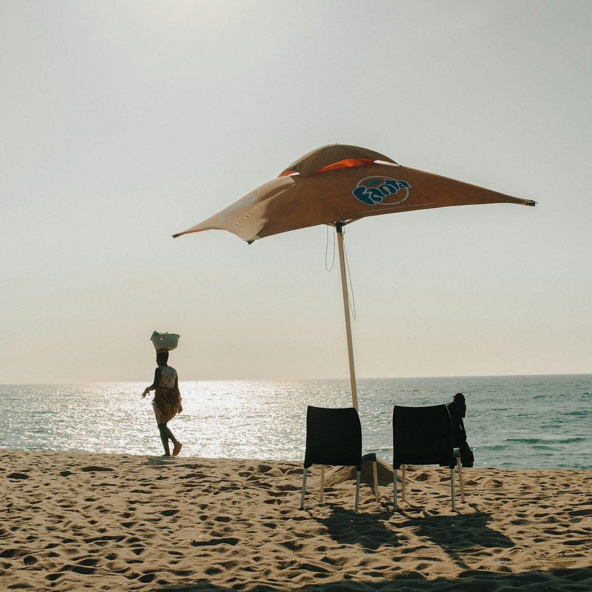 Woman walks on beach near umbrella and chairs.
