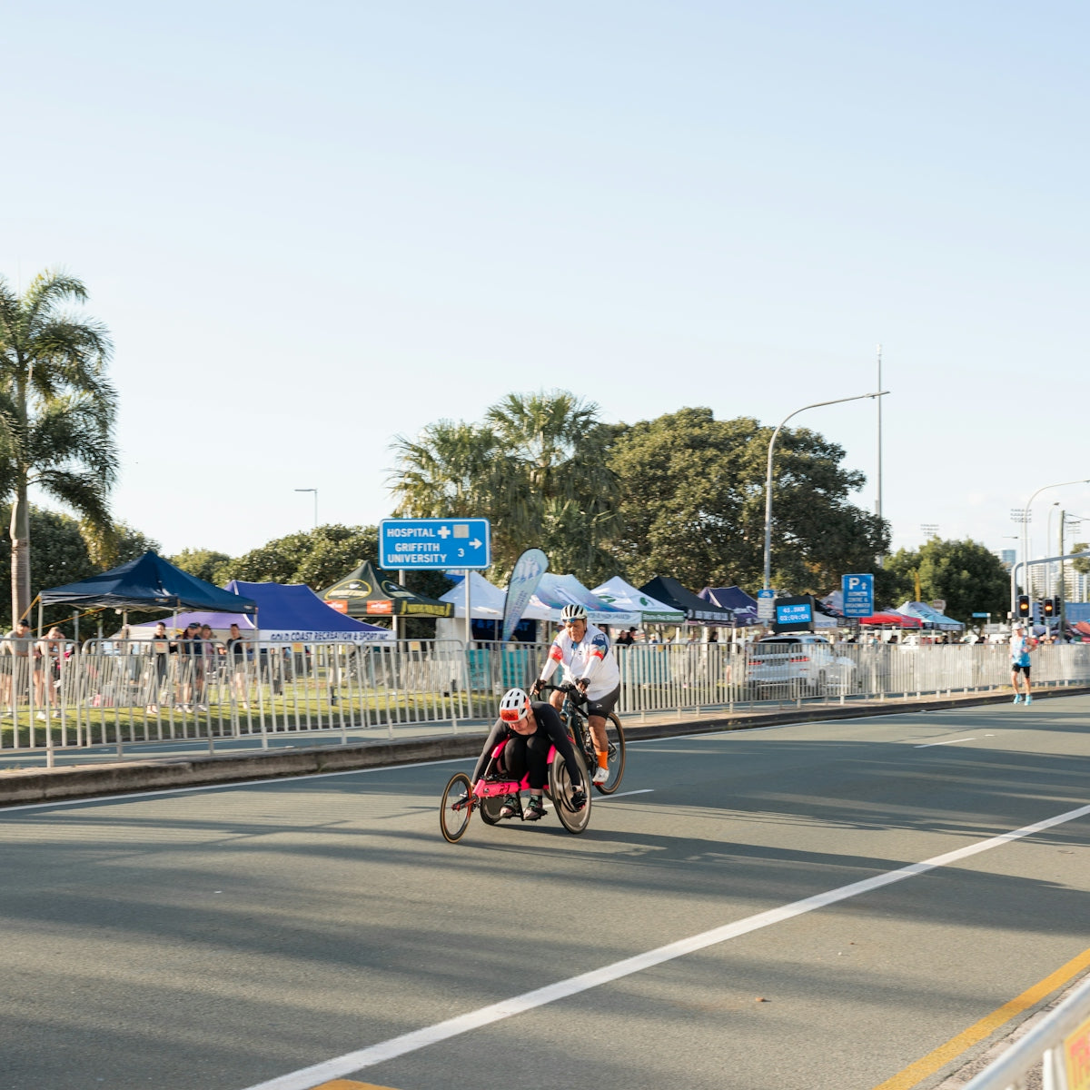 Handcyclist and cyclist on a road with event tents.